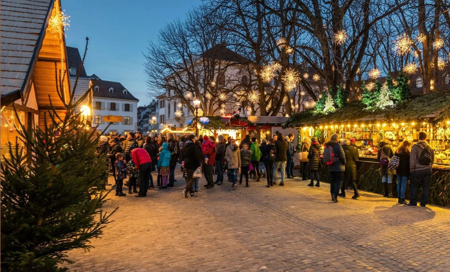 Mercado de Natal em Basileia, Suíça, com barraquinhas de madeira, luzes e o cenário histórico da Münsterplatz ao fundo.